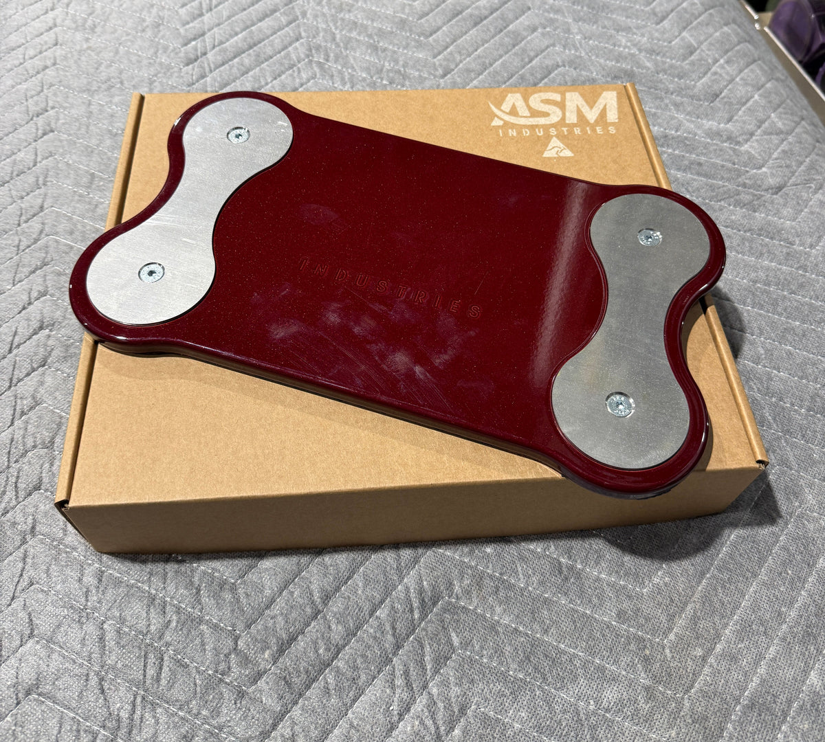 A dark red and silver ASM fitness plate resting on its brown cardboard box, placed on a gray quilted blanket.